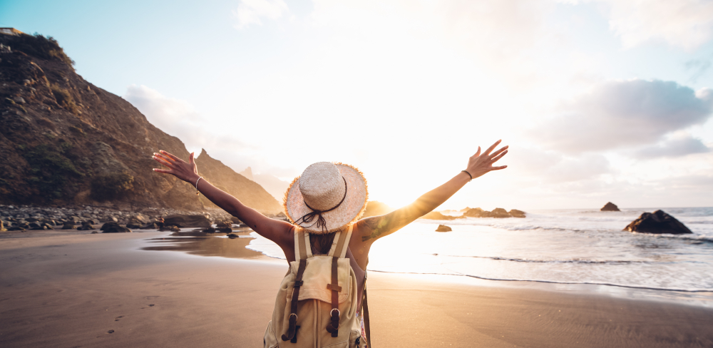 The meaning of life illustrated by a young backpacker on the beach, complete with sunhat, joyfully welcoming the sunrise above the sea.